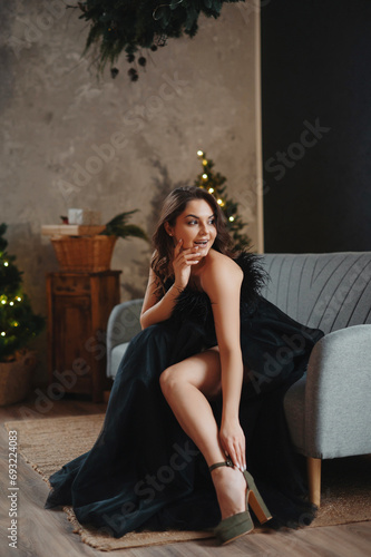 Happy cheerful young woman with curly brunette hair in evening dress sitting in armchair against Christmas tree. New year celebration concept