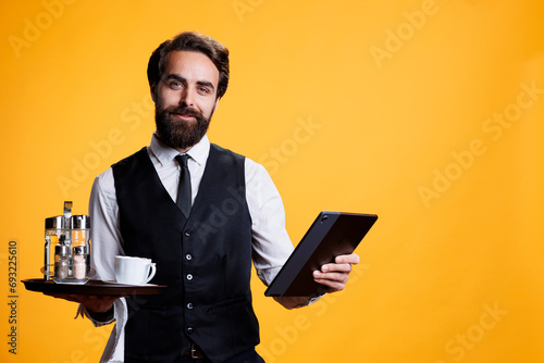 Experienced staff prepares bill on tablet, working at five star restaurant to serve people and provide excellent luxurious service. Butler in suit and tie carrying tray and modern gadget.