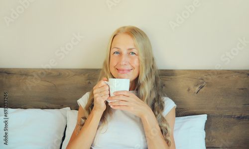 Happy smiling middle aged woman enjoying with cup of hot coffee lying on bed at home in the morning