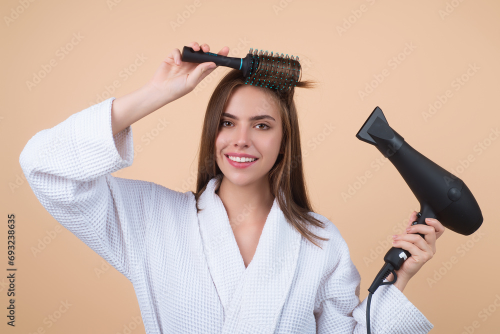 Woman in bathrobe combing hair, drying hair with hairdryer. Portrait of female model with a comb brushing hair. Girl with hairs brush and blow dryer. Hair care and beauty. Morning routine. - obrazy, fototapety, plakaty 