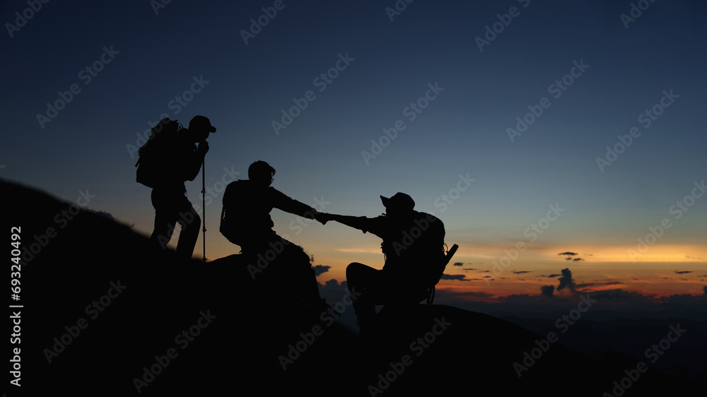 Silhouette of Asian teamwork hikers climbing up mountain cliff and one of them giving helping hand with friend at sunset, people helping, team work concept.