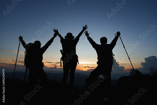 Silhouette of Asian three people standing raised hands with trekking poles and kerosene black lamp on cliff edge on top of rock mountain with at sunset rays over the clouds background,