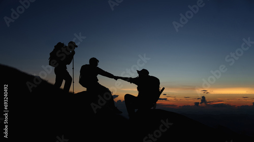 Silhouette of Asian teamwork hikers climbing up mountain cliff and one of them giving helping hand with friend at sunset, people helping, team work concept.