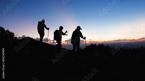 Silhouette of Asian teamwork hikers climbing up mountain cliff and one of them giving helping hand with friend at sunset, people helping, team work concept.