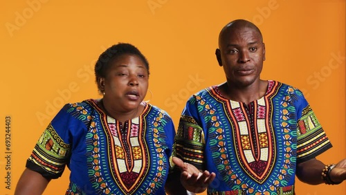 Unsure african american people doing i dont know sign in studio, showing doubtful clueless gesture on camera. Man and woman being uncertain, posing over orange background. photo