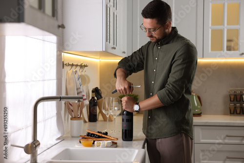 Romantic dinner. Man opening wine bottle with corkscrew at countertop in kitchen
