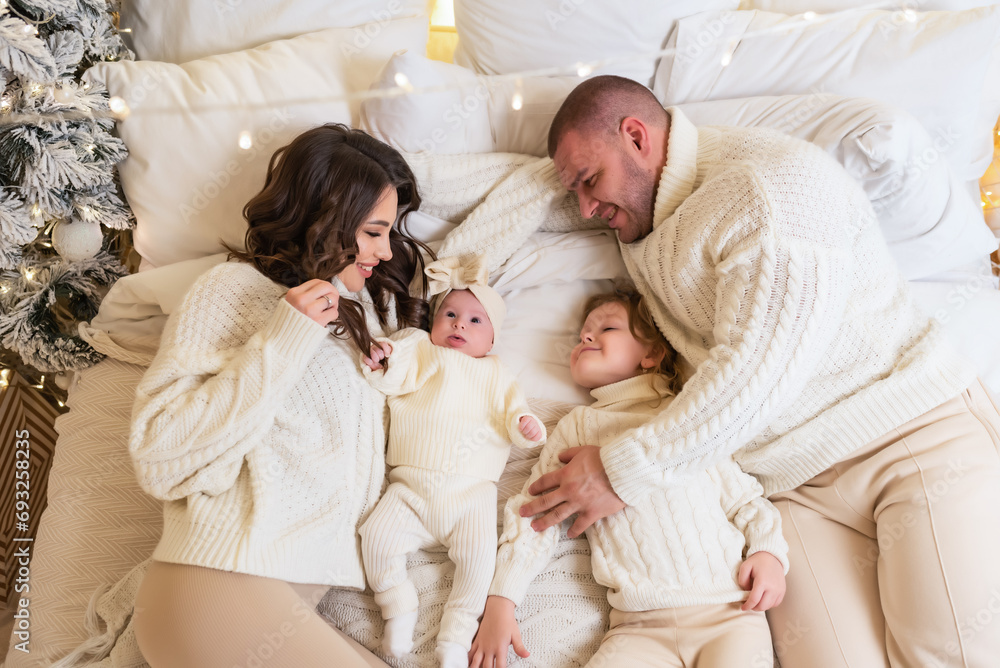 Young happy family wearing matching sweaters lies on the bed on Christmas morning. View from above. Father, mother and two daughters in a cozy bedroom decorated with a Christmas tree