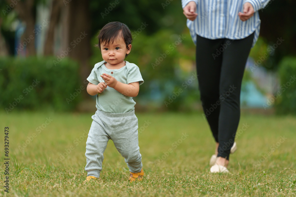 infant baby learn walking first step on grass field with mother helping in park infant baby learn walking first step on grass field with mother helping in park
