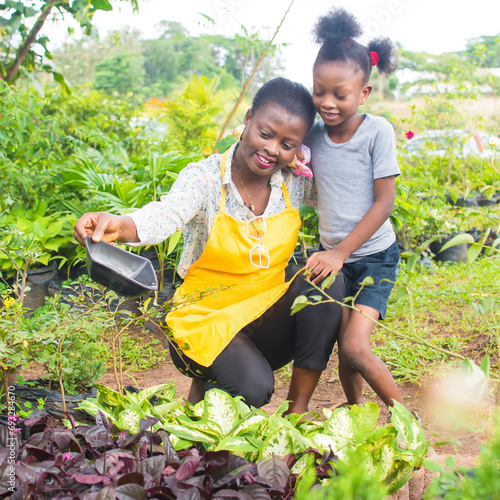 An African Nigerian female woman, mother, florist, entrepreneur, business lady in yellow apron, with a girl child beside her in an Horticulture garden filled with colorful green flowers in Nigeria