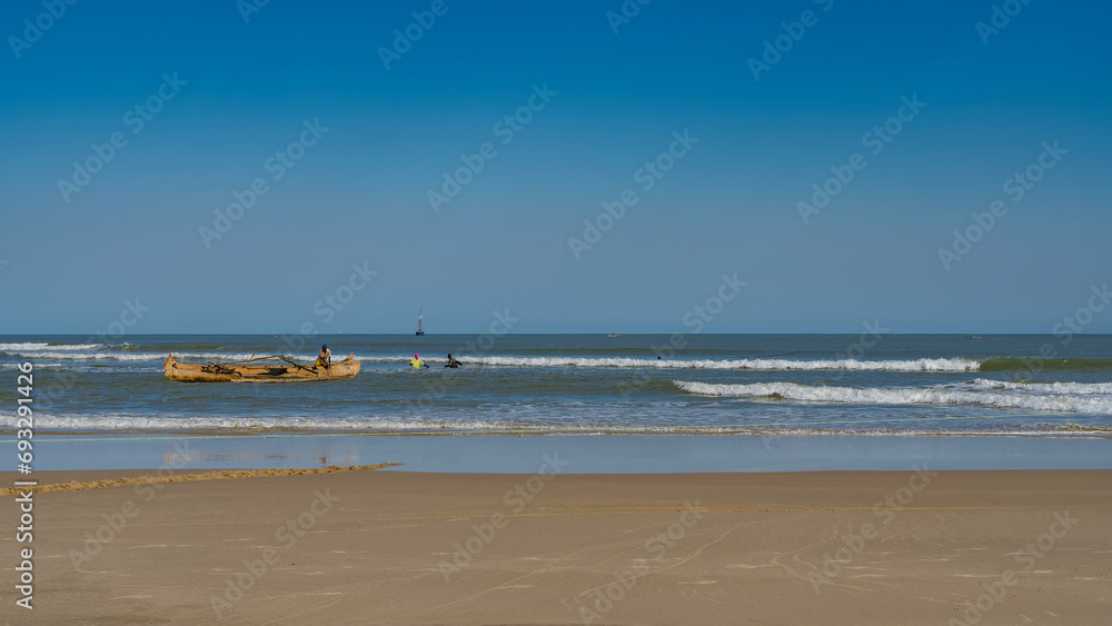 A wooden pirogue boat in the turquoise ocean. People in the water are pulling a fishing net. The waves are foaming and spreading over the sandy beach. Clear blue sky. Madagascar. Morondava. 