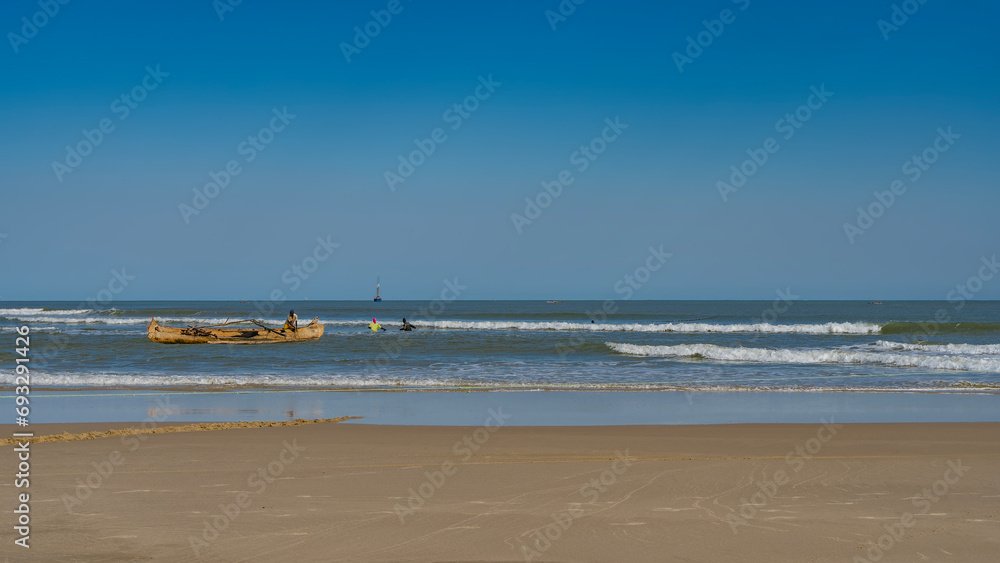 A wooden pirogue boat in the turquoise ocean. People in the water are pulling a fishing net. The waves are foaming and spreading over the sandy beach. Clear blue sky. Madagascar. Morondava. A wooden pirogue boat in the turquoise ocean. People in the water are pulling a fishing net. The waves are foaming and spreading over the sandy beach. Clear blue sky. Madagascar. Morondava.