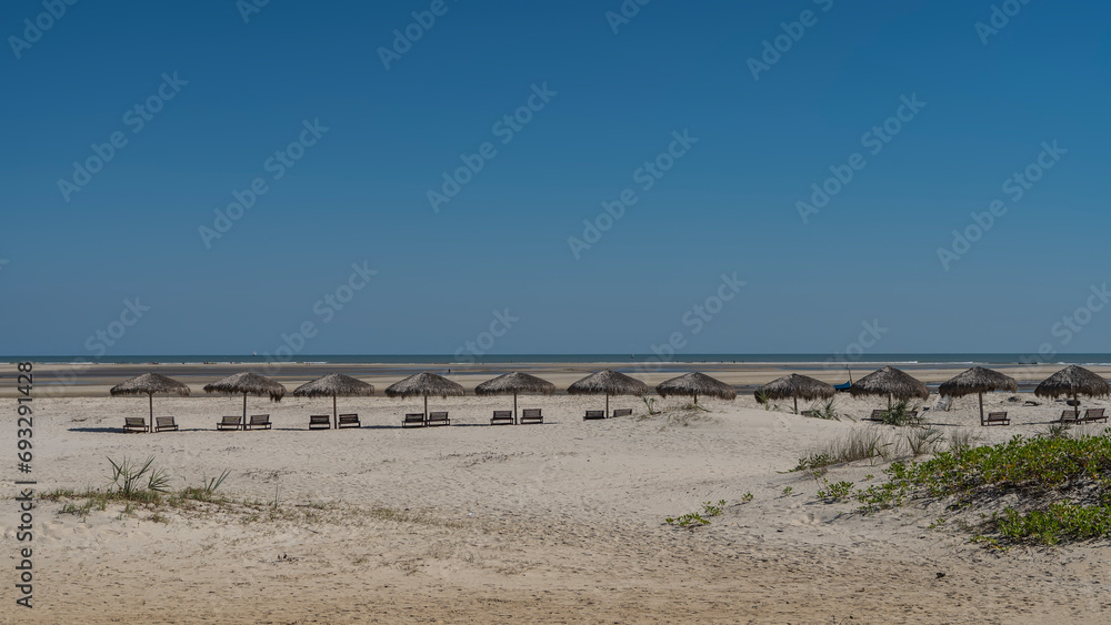 Sunbeds in the shade of straw sun umbrellas stand in a row on a wide sandy beach. Ahead, on the ocean shore, tiny silhouettes of people are visible. Madagascar. Morondava. The Mozambique Channel.