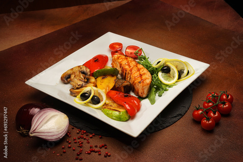 Baked salmon on a white plate with baked vegetables, served with red pepper, zucchini, mushrooms, herbs, tomatoes, onions, olives and lemon.