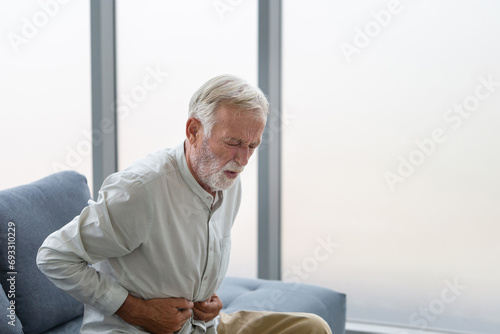 Unhappy senior man with stomach ache, man presses his hand to stomach from unbearable pain, mature man with stomach pain feeling unwell sitting in living room photo