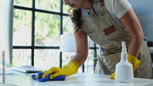 The young housekeeper wore gloves and cleaned the work table with a rag and cleaning spray. Professional office home cleaning products Providing hygiene services Free from germs.
