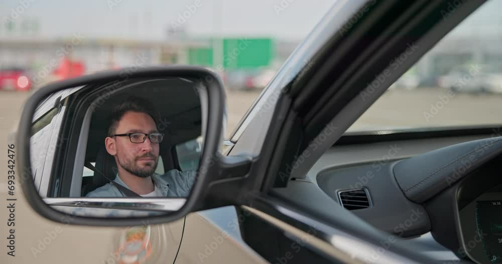 Reflection in car side view mirror - Young Caucasian man sits in a car on a parking lot and looking forward Reflection in car side view mirror - Young Caucasian man sits in a car on a parking lot and looking forward