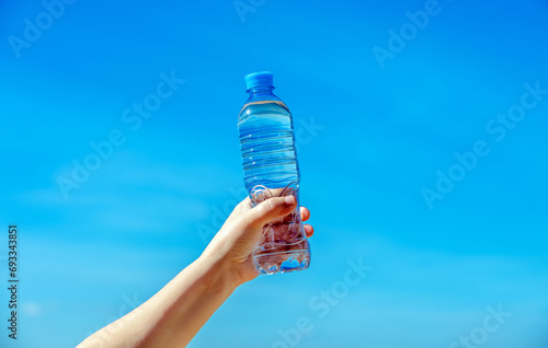 A girl holds a bottle of drinking water in her hand against a blue sky background 