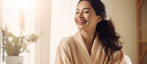 Happy young Asian woman in silk bathrobe enjoying morning beauty routine at home, applying face care product and smiling, with space for copying.