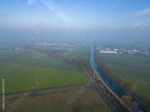 The image offers a bird's-eye view of a serene rural landscape shrouded in mist. In the foreground, a canal cuts a clear, straight path between fields of varying shades of green, hinting at different