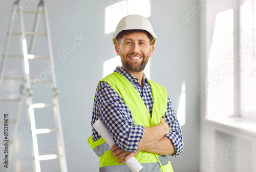 Portrait of smiling builder or architect in hardhat inside new house. Happy bearded man in hard hat and workwear standing with arms folded at home construction site with step ladder in background
