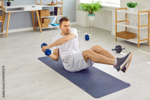 Fit, strong, young man sitting on a sports workout mat at home and doing an active fitness exercise with dumbbells. Physical activity, body training, strength concept