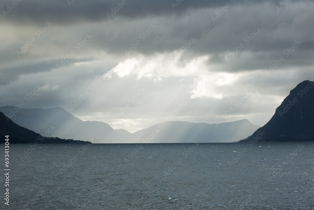 A body of water with mountains and sun striking through the cloudsin the background