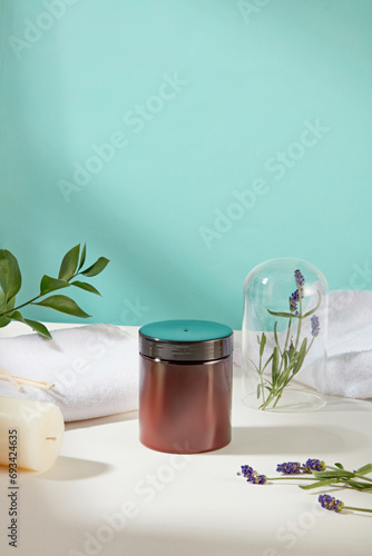 A brown jar of scrubs is displayed on the table with cotton towels, green leaves, a candle and lavender flowers. Skin care with products containing natural ingredients