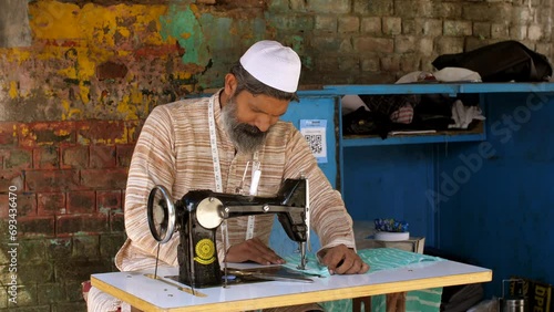 Indian muslim man making a dress on sewing machine - tailoring  man with skull cap  low-wage worker  self-employed. An Indian tailor sews the cloth using hand-powered machine - dressmaker  tailor  ...