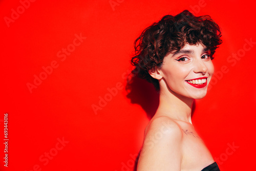 Fashion beauty portrait of young smiling brunette woman with evening stylish  makeup and perfect clean skin. Sexy model with curly short hair posing in studio. With red bright lips. Isolated on red