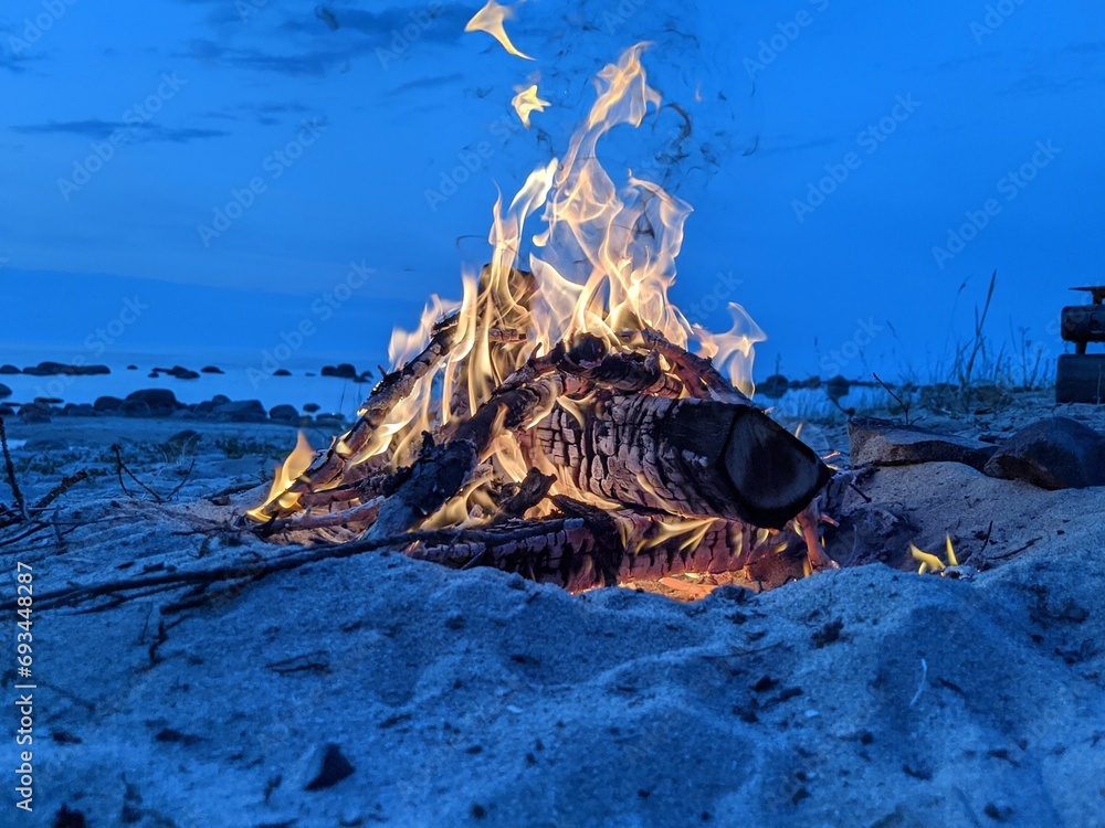 Blazing campfire on the beach during summer evening. Bonfire in nature as background. Burning wood on white sand shore at sunset. selective focus. tropical romantic landscape near sea water edge.