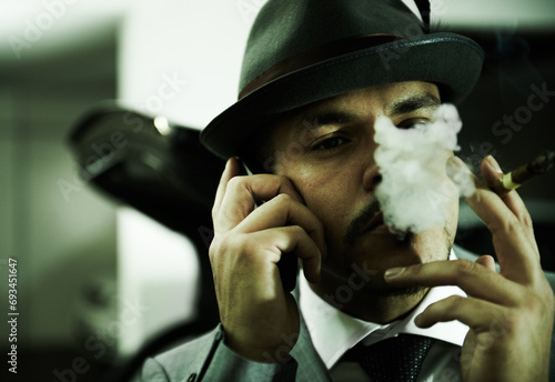 Man, gangster and phone call while smoking or planning mafia, danger or ransom abduction. Male person, cigar and mobile device or hitman conversation for robbery violence, interrogation or terror photo