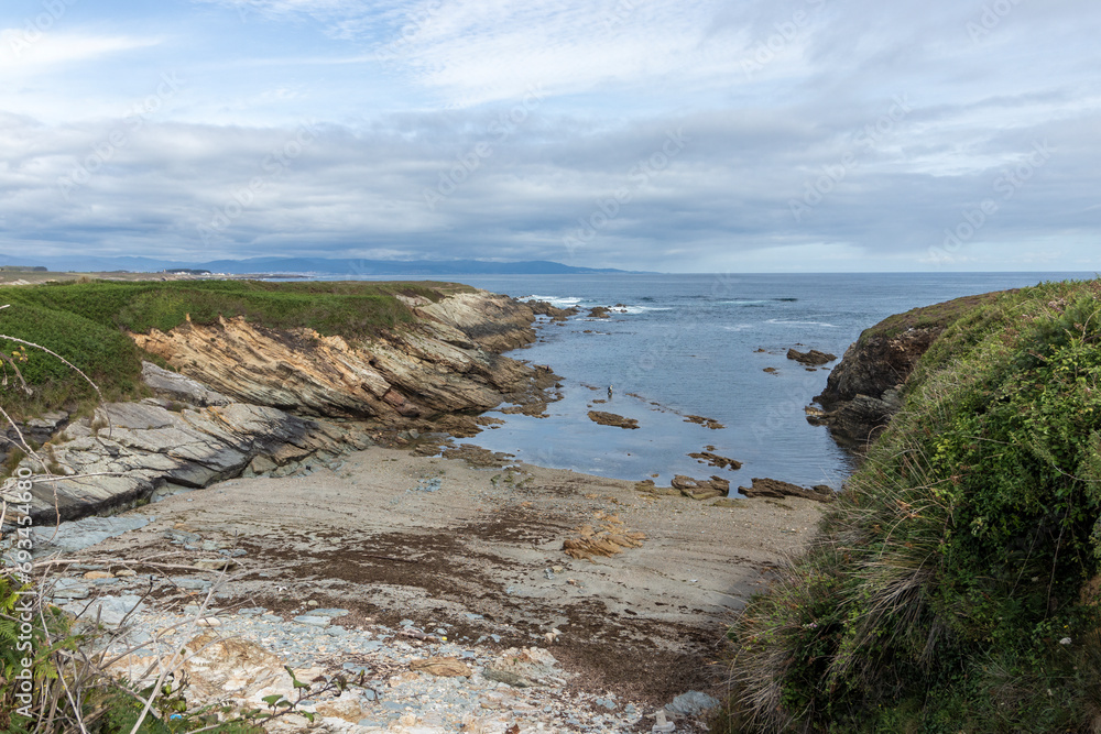 A serene coastal landscape with a rocky shoreline, greenery, calm sea, and distant mountains under a cloudy sky