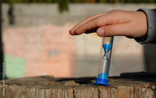 Child's hand with sandglass or sand timer in the home yard. Wall background. Sunlight. Copy space. 