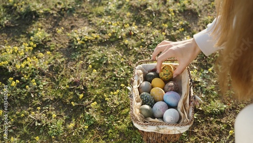 A painted Easter egg lies in a clearing among yellow flowers. The young woman picks it up and puts it in a basket with other eggs. close-up photo
