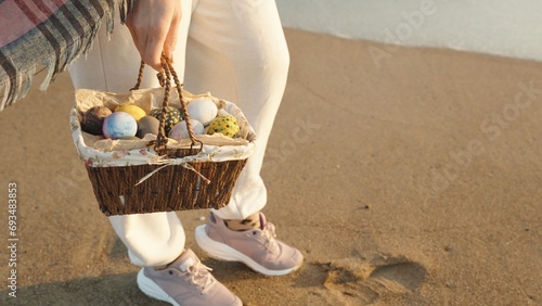 I'm standing on the sand by the sea and holding a wicker brown basket with Painted Easter Eggs.