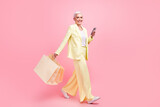 Full size photo of cheerful woman dressed yellow suit go to empty space holding shopping bags smartphone isolated on pink color background