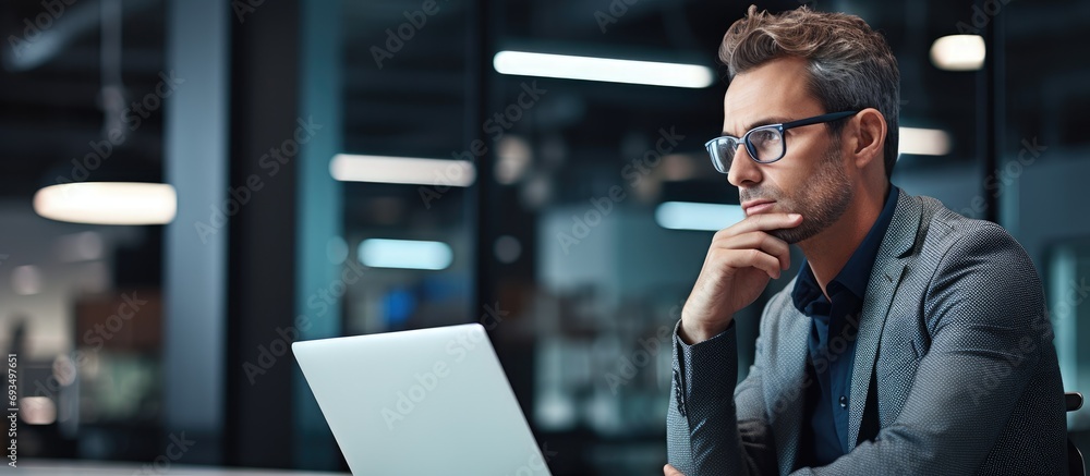 Content businessman using laptop in office, looking away. Pensive professional considering AI solutions. - obrazy, fototapety, plakaty 