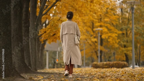 back view of an attractive brunette woman wearing coat walking in park at autumn day with yellow dry leaves on trees. happy beautiful woman in autumn outdoors. Slow-motion 4k footage