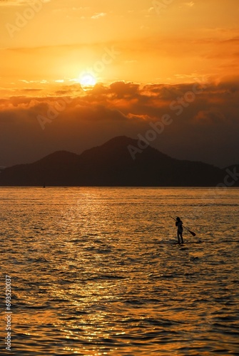 Silhouette of woman practicing stand up paddle boarding in the sea with mountains in the background during sunset.