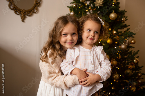 Two sisters play near Christmas tree indoors. Merry Christmas and Happy Holidays.