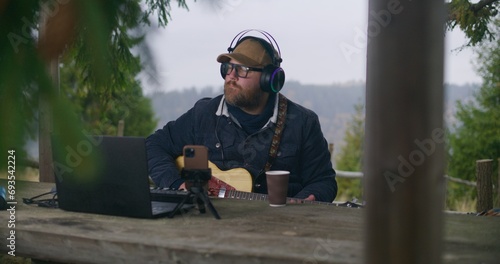 Caucasian man in headphones plays guitar sitting in wooden gazebo in the forest. Male musician composes and records music using laptop and phone on tripod during vacation trip in the mountains. photo