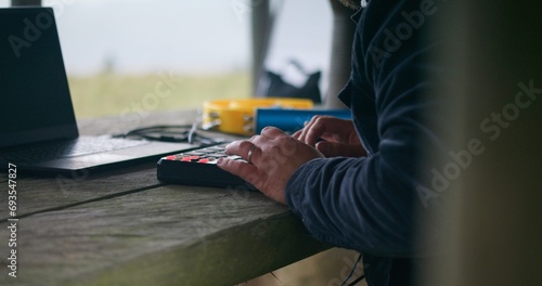 Close up of Caucasian musician in headphones playing digital electric piano, moving rhythmically and creating music sitting in gazebo. Man uses laptop to record song during vacation in the mountains.