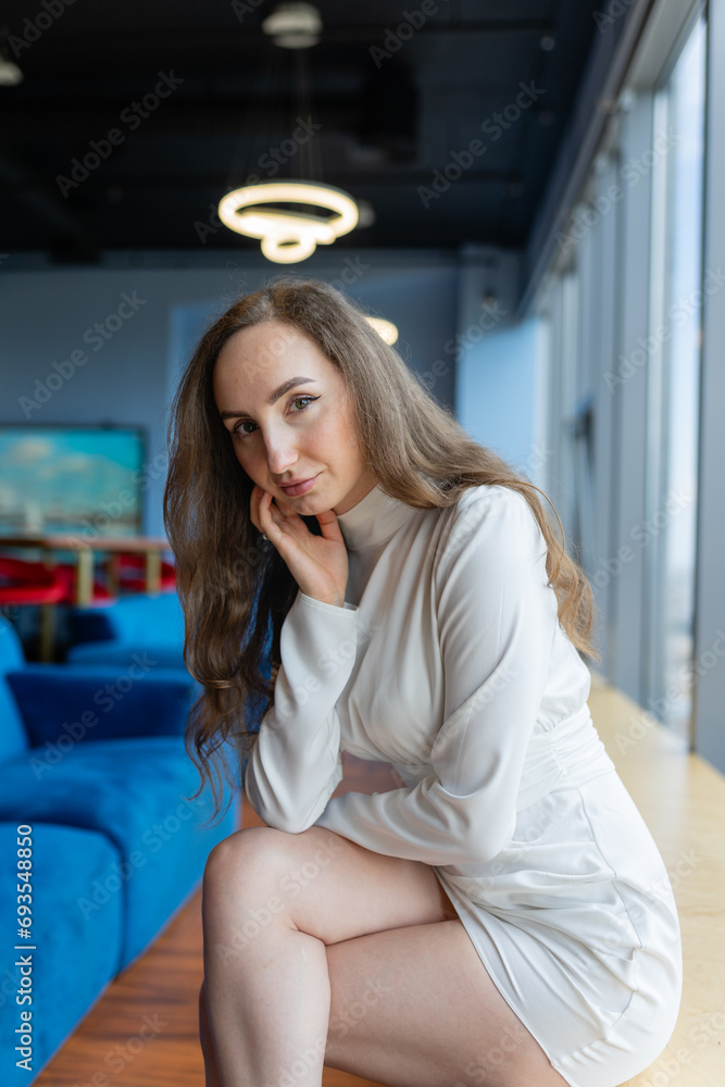 Businesswoman sits on wooden bench near windows in company lounge zone. Executive manager in short dress with bare legs near windows