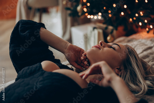 Young dreamy woman lying down on bed in black decollete dress near Christmas tree.