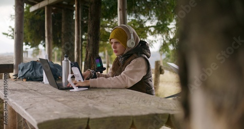 Teenager in headphones uses laptop and phone on tripod to create content for viewers during holidays in mountain forest. Young streamer sits in wooden gazebo and talks into professional microphone.