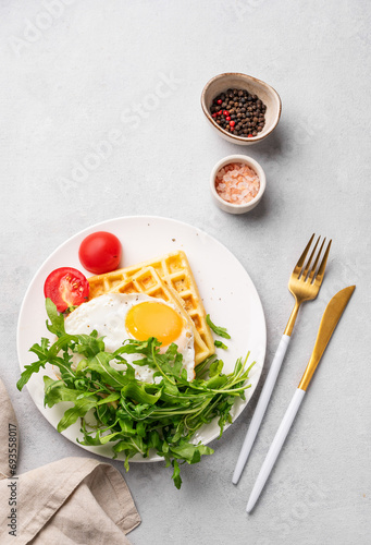 Waffles with fried egg, cherry tomatoes and arugula on a white plate on a gray texture background with spices.