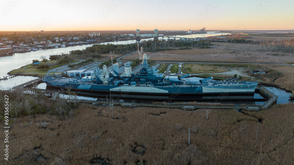 Battleship at sunrise in Wilmington, North Carolina, with the Cape Fear River flowing on the left and the Cape Fear Memorial Bridge in the distance, set against a backdrop of the awakening city.