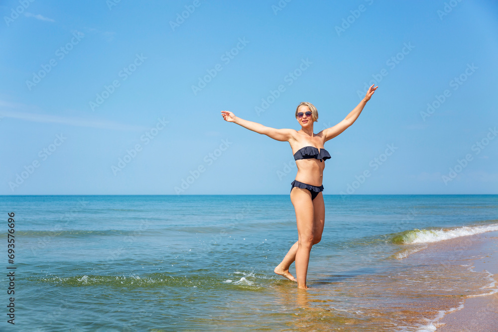 Laughing woman in a black bikini on the seashore. Active lifestyle, travel and relaxation at the resort on a sunny day. Full height.