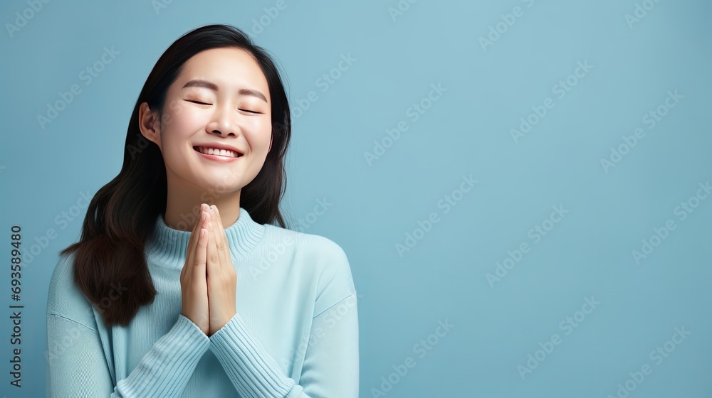 A beautiful asian woman standing against a blue background smiles with crossed fingers and looks hopeful while wishing for good luck. A beautiful asian woman standing against a blue background smiles with crossed fingers and looks hopeful while wishing for good luck.