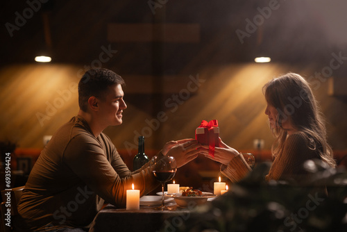 Happy young couple celebrating Valentines day having romantic dinner at home. Loving man giving gift box his woman